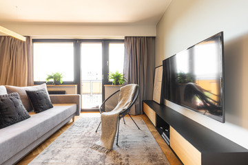 Blanket on black armchair between cupboard and grey sofa in bright living room interior. Real photo