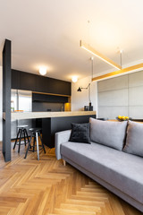 Pillows on grey sofa next to black kitchen with lights in modern living room interior. Real photo