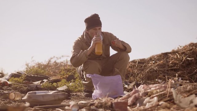 portrait of a dirty homeless hungry man in a dump drinks the missing juice in the package with walking goes looking for food slow motion video. homeless dirty man roofless person looking for food in a