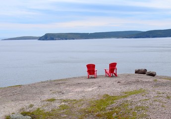 two empty red Adirondack chairs on the edge of a cliff overlooking the ocean and coastline