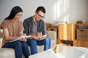Smiling Asian attractive female and handsome male in glasses sitting in earphones on sofa and writing down in notepads information from tablet placed on desk nearby .
