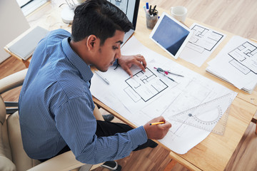 From above adult Indian architect working on blueprint while sitting at desk in office