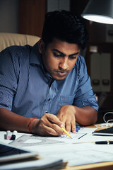 Adult Indian employee sitting in office and overtiming on blueprint