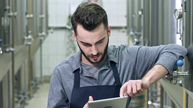 Handsome brewery worker leaning on beer vessel at plant and surfing the Internet on digital tablet