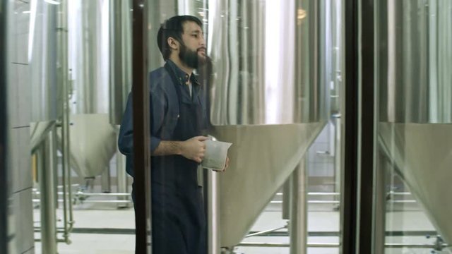 View through the glass wall of brewery technician in apron holding grist in plastic container and walking through steel vessels