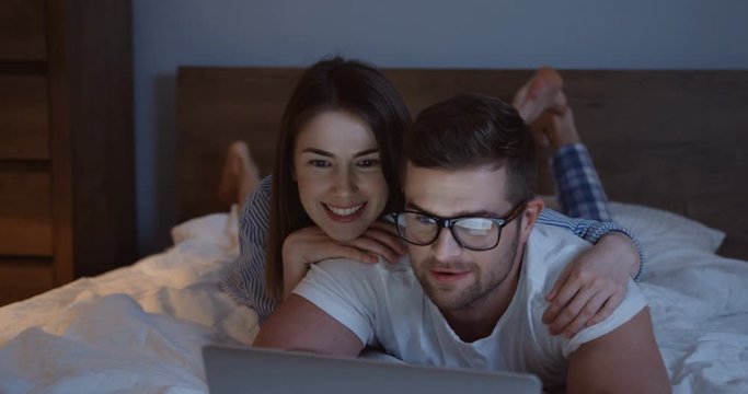 Close up of the young attractive couple lying on the bed in the evening and watching something funny on the laptop computer.