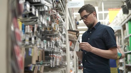Master is choosing tool for a finishing works and tiling in a shop for builders. Man is looking on a toothed spatula, standing near rack
