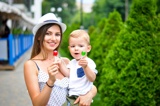 A young beautiful mother in a white hat is holding a two-year-old son and they are eating colored lollipops.