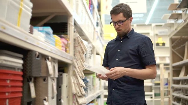 Male shopper is choosing plastic containers for storage in a hypermarket. He is opening, twirling in hands and collating sizes