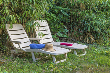 Holiday concept: two white lounge chairs, towels, straw hat in greenery of bamboo
