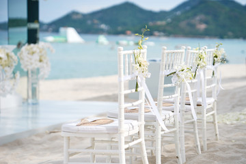 Side view of white chiavari chairs arrangement for beach wedding venue, flower with white ribbin decoration on back side. Panoramic sea view and small island background