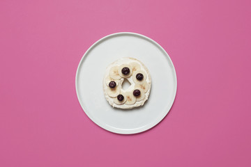 Blueberry and banana bagel with cream cheese on a white plate on a pastel pink background