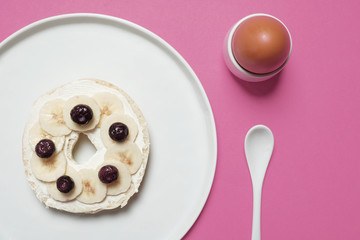 Blueberry and banana bagel with cream cheese on a white plate on a pastel pink background with a soft boiled egg and a white spoon