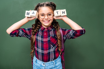 smiling schoolchild holding wooden cubes with word math near blackboard