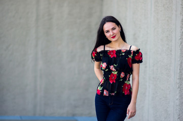 The young girl in black top with red flowers and open shoulders stands on the gray wall background and smiles with red lips