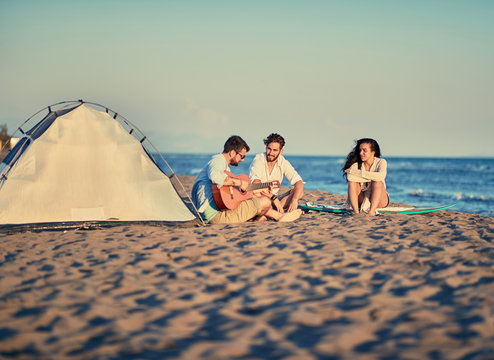 summer, vacation, holiday, happy people concept - friends relaxing with playing guitar and sing a song on sea beach.