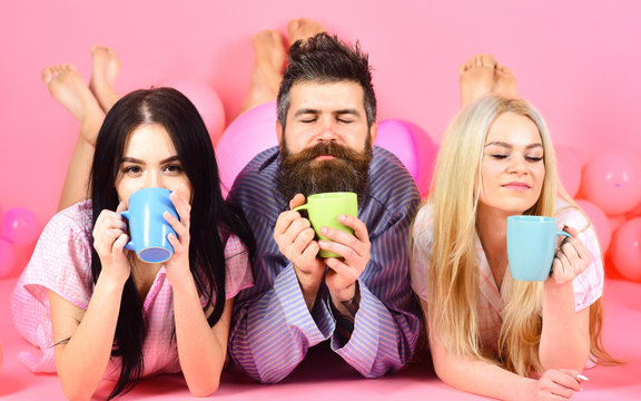 Threesome relax in morning with coffee. Lovers concept. Man and women, friends on sleepy faces lay, pink background. Man and women in domestic clothes, pajamas. Lovers in love drink coffee in bed.