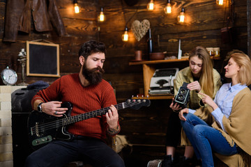 Fototapeta na wymiar Man with long beard enjoying music. Musician with calm, concentrated face playing for his wife and daughter. Mom helping teenage girl to deal with retro camera. Bearded man tuning electrical guitar