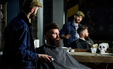 Obraz premium Hipster client got new haircut. Barber with hair clipper looking at mirror, barbershop background. Professional master checking result while client sits in chair. Barber done haircut. Haircut concept