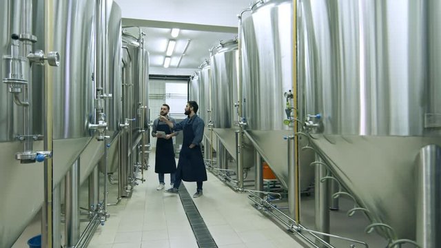 Two coworkers walking through beer factory between rows of steel brewing vessels, using digital tablet, inspecting equipment and talking