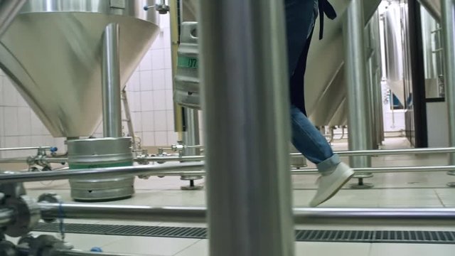 Tracking shot of brewery worker carrying beer keg along row of steel brewing vats