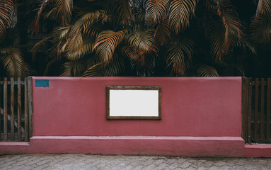 Blank information banner on the pink concrete fence with tropical trees behind; empty mock-up of the signboard on the fencing; white clean billboard in old wooden frame with palm branches behind