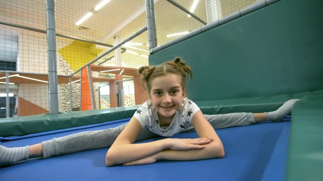 Medium shot of adorable little girl sitting in middle split on indoor trampoline and looking at camera