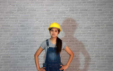 Technician woman ware white helmet with grey T-shirt and denim jeans apron dress standing akimbo on grey brick pattern background.