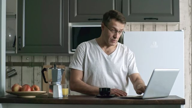 Medium shot of middle-aged freelance worker opening laptop computer and pouring coffee into cup when beginning work at home in the morning after waking up