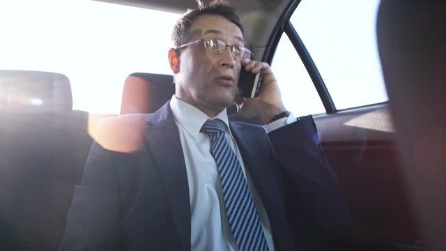 Medium shot of middle-aged male entrepreneur in business suit and glasses talking on phone in backseat of taxi when going to work in the morning