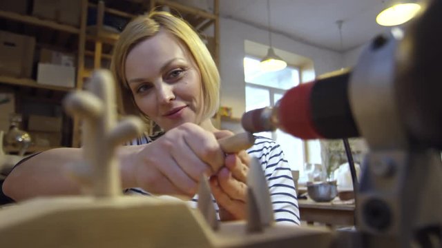 Medium shot of concentrated blonde woman polishing small wooden detail using sanding band attached to drill