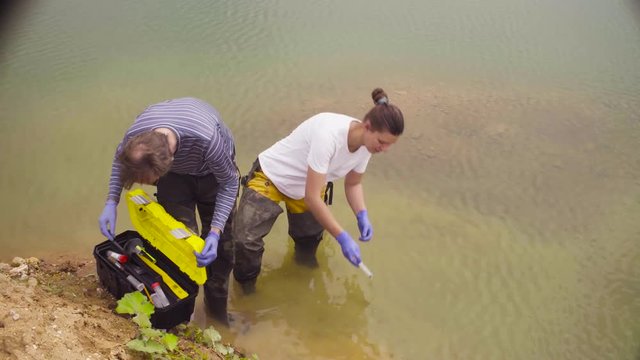 Man and woman scientist environmentalist standing in a river. Woman taking sample of water and pouring it into the test tube.