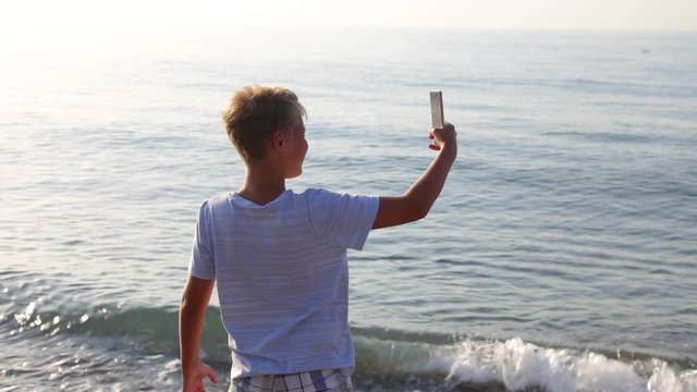 Back view of cute funny blonde boy making selfie on sea beach at sunny sunrise sea and sky background. Happy beach holiday concept. Real time full hd video footage.