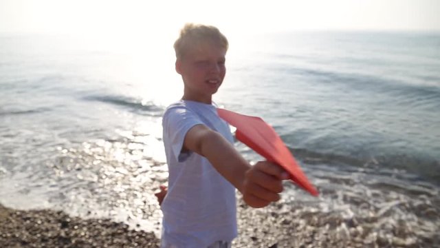 Closeup portrait of happy smiling white child playing paper airplane at sunny blue sunset sea water background on early summer morning. Real time full hd wide angle video footage.
