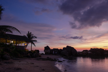 Beach, rocks and beach bar shadowed under the palm trees on the background of the sunset almost...