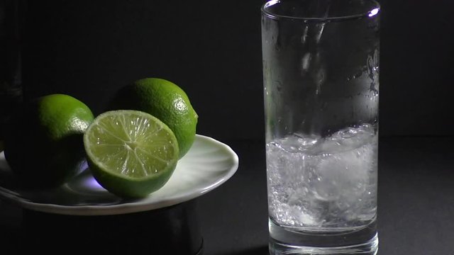 In a glass of ice poured soda water, next to the lime fruit on a white plate on a black background