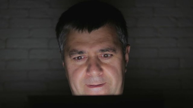 A strange man with the appearance of a maniac looks at the screen of the tablet in a dark room.