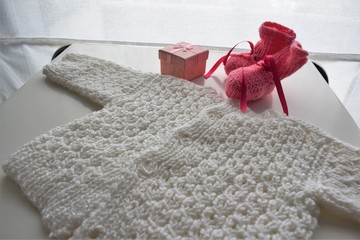 Gifts for new born baby girl in winter: a pair of  pink wool shoes, a handmade woven white pullover and a golden gift box.