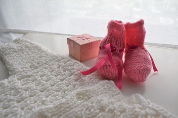 Gifts for new born baby girl in winter: a pair of  pink wool shoes, a handmade woven white pullover and a golden gift box.