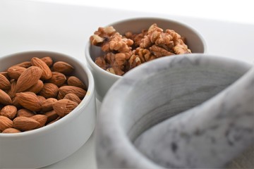 Three white containers with almonds, walnuts and a mixture of sunflower, sesame and chia seeds near a marble mortar.