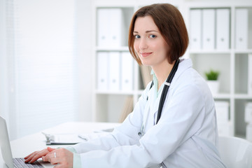 Young brunette female doctor typing on laptop comoputer while sitting at the table in  hospital office.  Health care, insurance and help concept