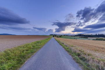 Countryside with harvested cereal field and paved laneway at dusk in summer at Roellbach in Spessart hills in Bavaria, Germany
