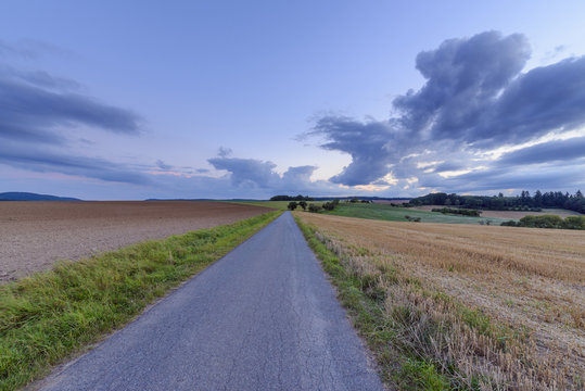 Countryside with harvested cereal field and paved laneway at dusk in summer at Roellbach in Spessart hills in Bavaria, Germany
