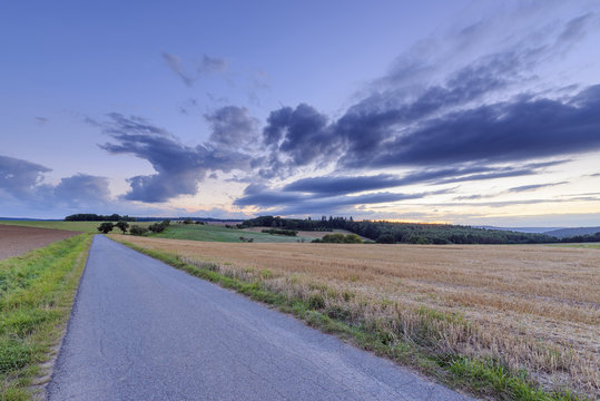 Countryside with harvested cereal field and paved laneway at dusk in summer at Roellbach in Spessart hills in Bavaria, Germany