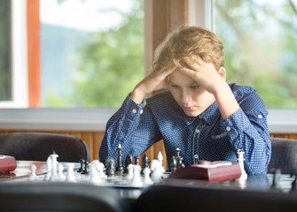 cute young smart boy in blue shirt plays chess on the training before the tournament. chess summer camp
