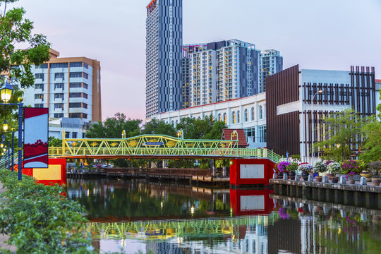 Footbriidge over river at dusk in Malacca Malaysia with city buildings in the background.