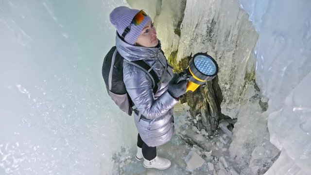Travel woman on ice cave of Lake Baikal. Trip to winter island. Girl backpacker is walking of ice grot. Traveler looks at beautiful ice grotto. Hiker wears sports glasses, silver jacket, backpack, led
