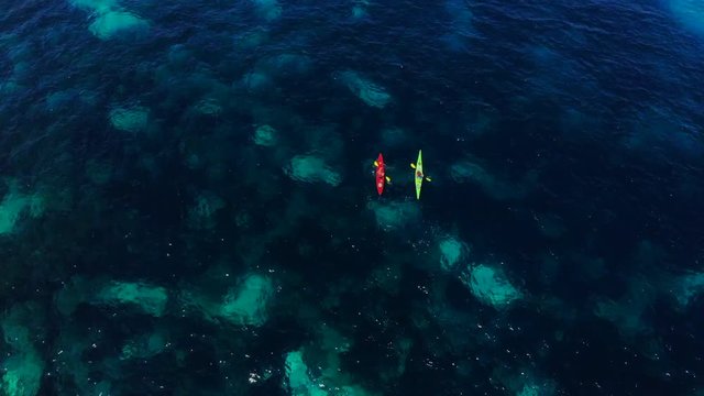 Flying over two kayaking people. The overhead shot emphasises the dark and light blue pattern of the sea below them and therefore gives the shot a tropical touch. PAN UP at the end to an island