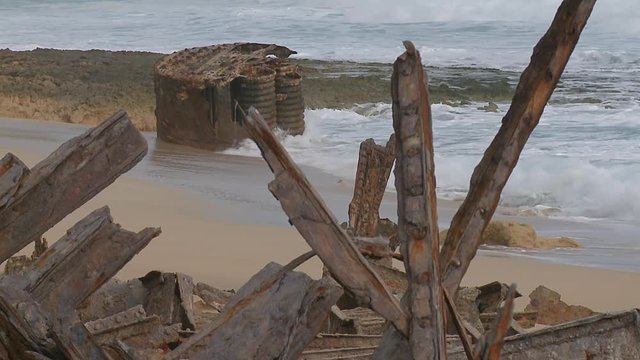 Remains of a stranded ship on a sandy beach in fore- and background surrounded by the surf