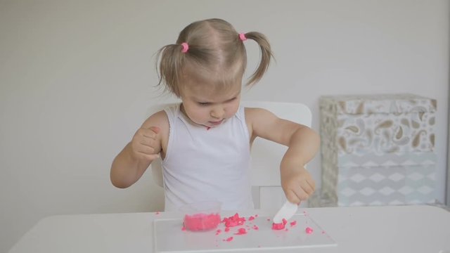 A little girl sitting at a table in a bright room sculpts a pink plasticine figurines